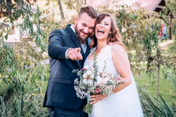 Novomanželský pár se šťastně směje, ženich ukazuje prstem, nevěsta drží kytici.