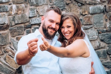 Novomanželský pár se směje a drží miniaturní prstýnek před kamennou zdí.