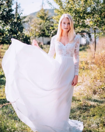 Žena v dlouhých bílých šatech s krajkovými rukávy stojí na slunné zahradě.