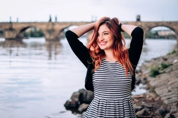 Žena s červenými vlasy a pruhovanými šaty stojící u řeky, v pozadí historický kamenný most.