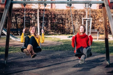 Dvě ženy sedí na houpačkách v parku, jedna v žlutém svetru a druhá v červeném.