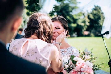 Nevěsta v krajkových šatech dojatá při svatebním obřadu, drží ji osoba v květovaných šatech.