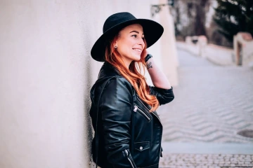 Žena s dlouhými červenými vlasy, v kožené bundě a klobouku, usmívající se opřená o bílou zeď.