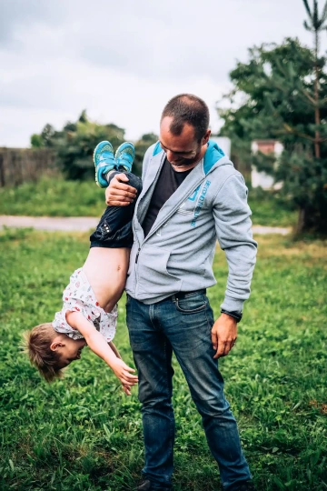 Muž v šedé mikině drží dítě za nohy vzhůru nohama na trávníku, oba se smějí.