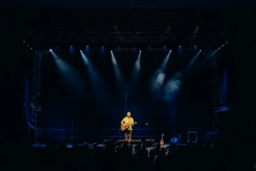 Muž hrající na kytaru na osvětleném pódiu před publikem.