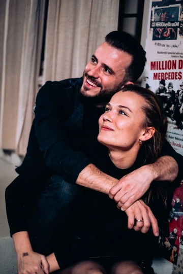 Pár sedící vedle sebe, muž objímá ženu, oba se usmívají a dívají se nahoru.