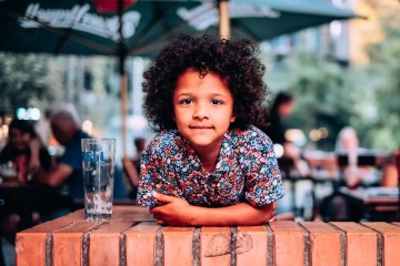 Dítě s kudrnatými vlasy v květované košili se opírá o cihlovou zeď venku.