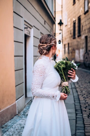 Nevěsta ve svatebních šatech s kyticí květin stojí v městské uličce.