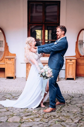 Svatební pár se směje, muž v obleku a žena v šatech drží kytici, dřevěný nábytek v pozadí.