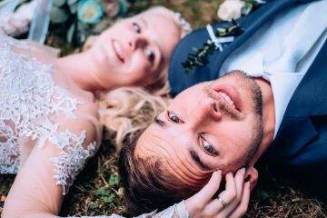 Nevěsta a ženich leží v trávě, nevěsta má krajkové šaty, ženich oblečený v tmavém obleku s bílou kravatou.