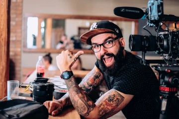 Lidé natáčející video v kavárně, muž s tetováním a čepicí ukazuje palcem na kameru.