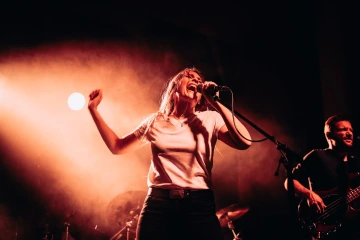 Zpěvačka energicky vystupující na koncertě s kytaristou v pozadí, osvětlena oranžovým reflektorem.