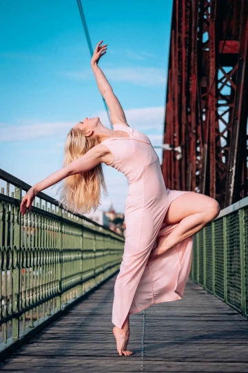 Tanečnice v růžových šatech balancuje na mostě pod modrou oblohou.