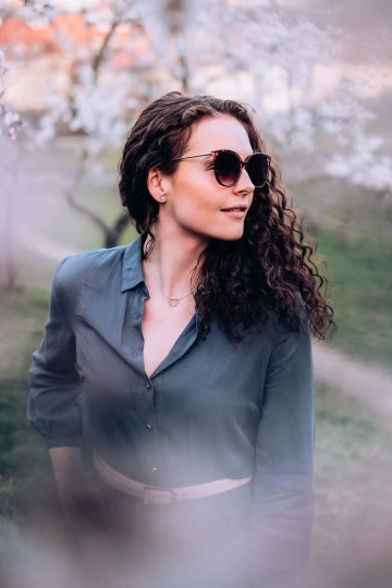 Žena s tmavými brýlemi a kudrnatými vlasy pózuje u rozkvetlého stromu.