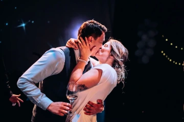 Pár se líbá na svatební oslavě, muž drží sklenku vína.