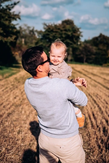 Muž v brýlích drží usmívající se dítě na polní cestě za slunečného dne.