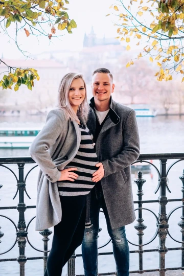 Těhotný pár v parkovém prostředí u řeky, muž jemně objímá břicho partnerky.