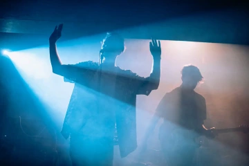 Zpěvák na pódiu zvedá ruce během koncertu, vedle něj hraje kytarista, obklopeni modrými světly a kouřem.