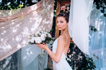 Nevěsta s kyticí bílých růží stojí u romantické látkové dekorace.
