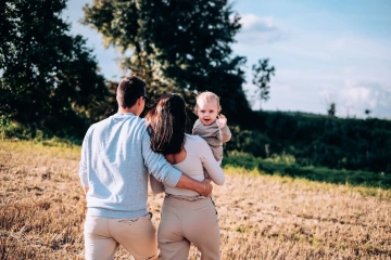 Rodina na procházce po poli, rodiče drží dítě, které mává.