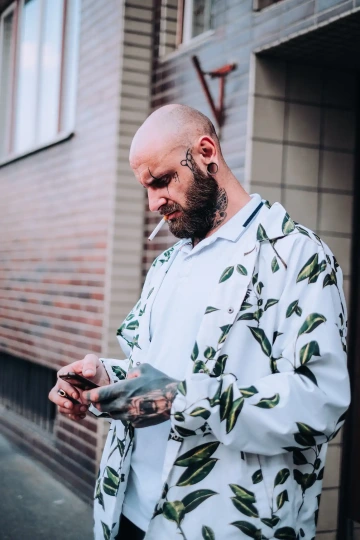 Tetovaný muž s cigaretou v ústech používá telefon, oblečený v bílé košili s listovým vzorem.
