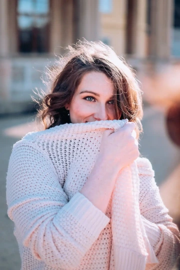 Žena s úsměvem zakrývající ústa pleteným svetrem, venku za jasného dne.