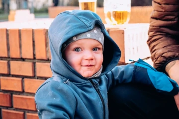Dítě v modré bundě a čepici s puntíky sedí venku u cihlové zdi, v pozadí sklenice piva.